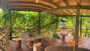 Una terraza de madera al aire libre con una mesa redonda y sillas, rodeada de abundante vegetación.