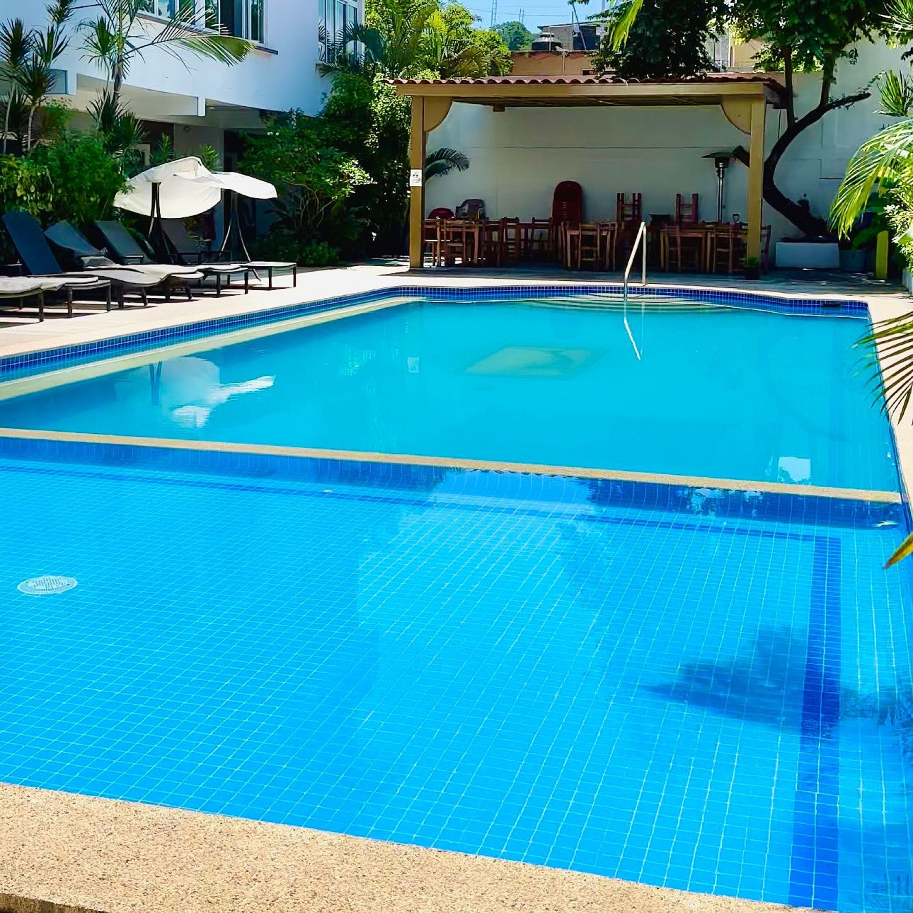 Una piscina rectangular con azulejos azules, rodeada de sillones de playa y sombrillas, y un área cubierta con mesas y sillas.