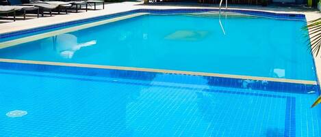 Una piscina rectangular con azulejos azules, rodeada de sillones de playa y sombrillas, y un área cubierta con mesas y sillas.