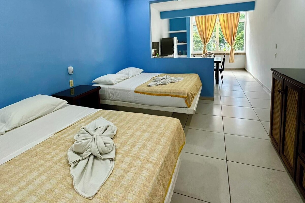 A hotel room with two beds, a ceiling fan, a window with curtains, and a kitchenette area.