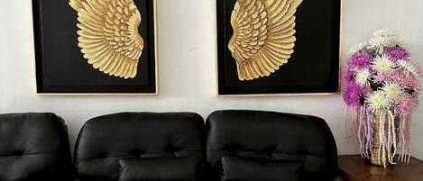 Ein Wohnzimmer mit einem schwarzen Ledersofa, zwei gerahmten goldenen Flügeln an der Wand und einer Blumenvase auf einem Beistelltisch.