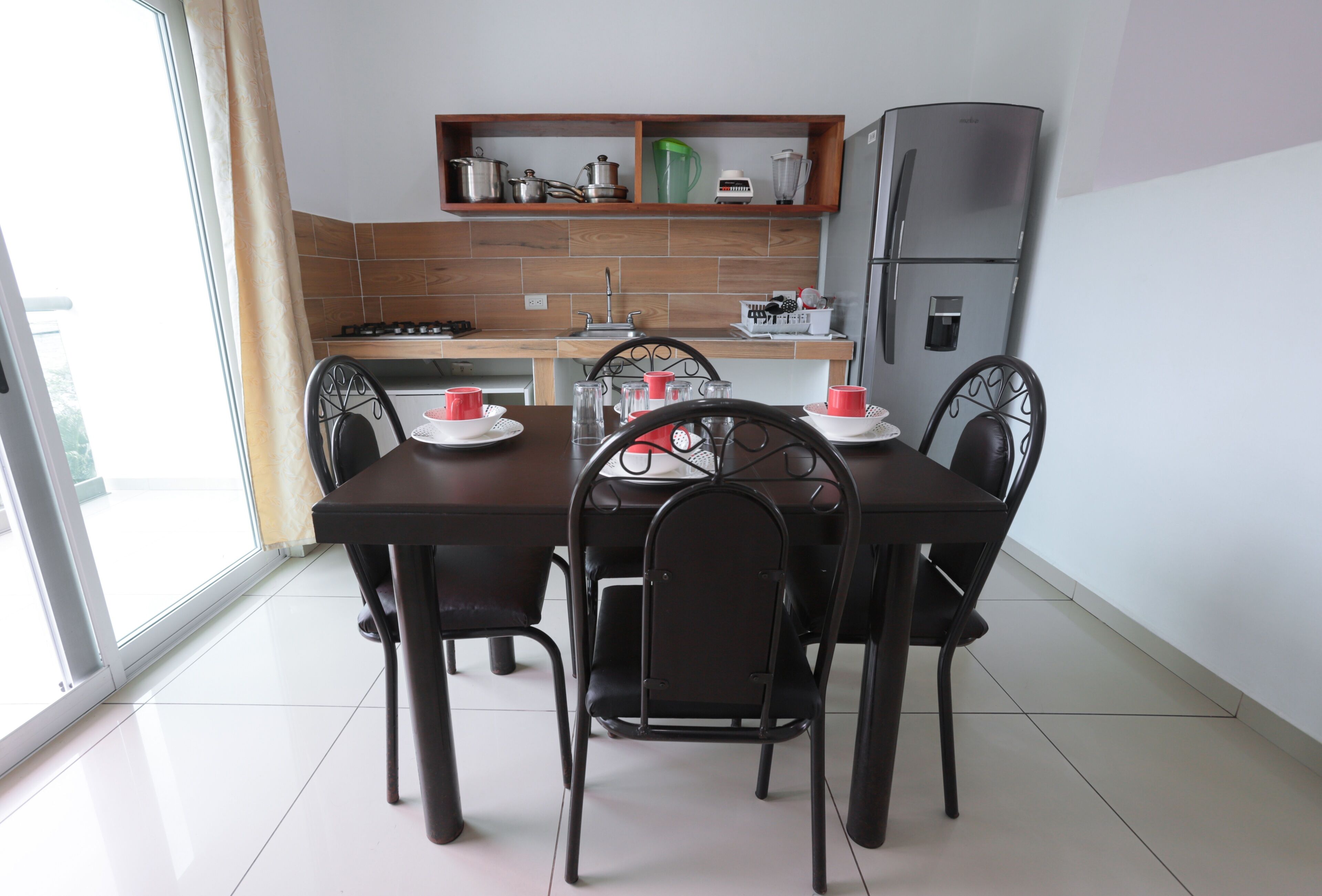 A dining area with a table and chairs, a kitchen with a refrigerator, and a window with a curtain.