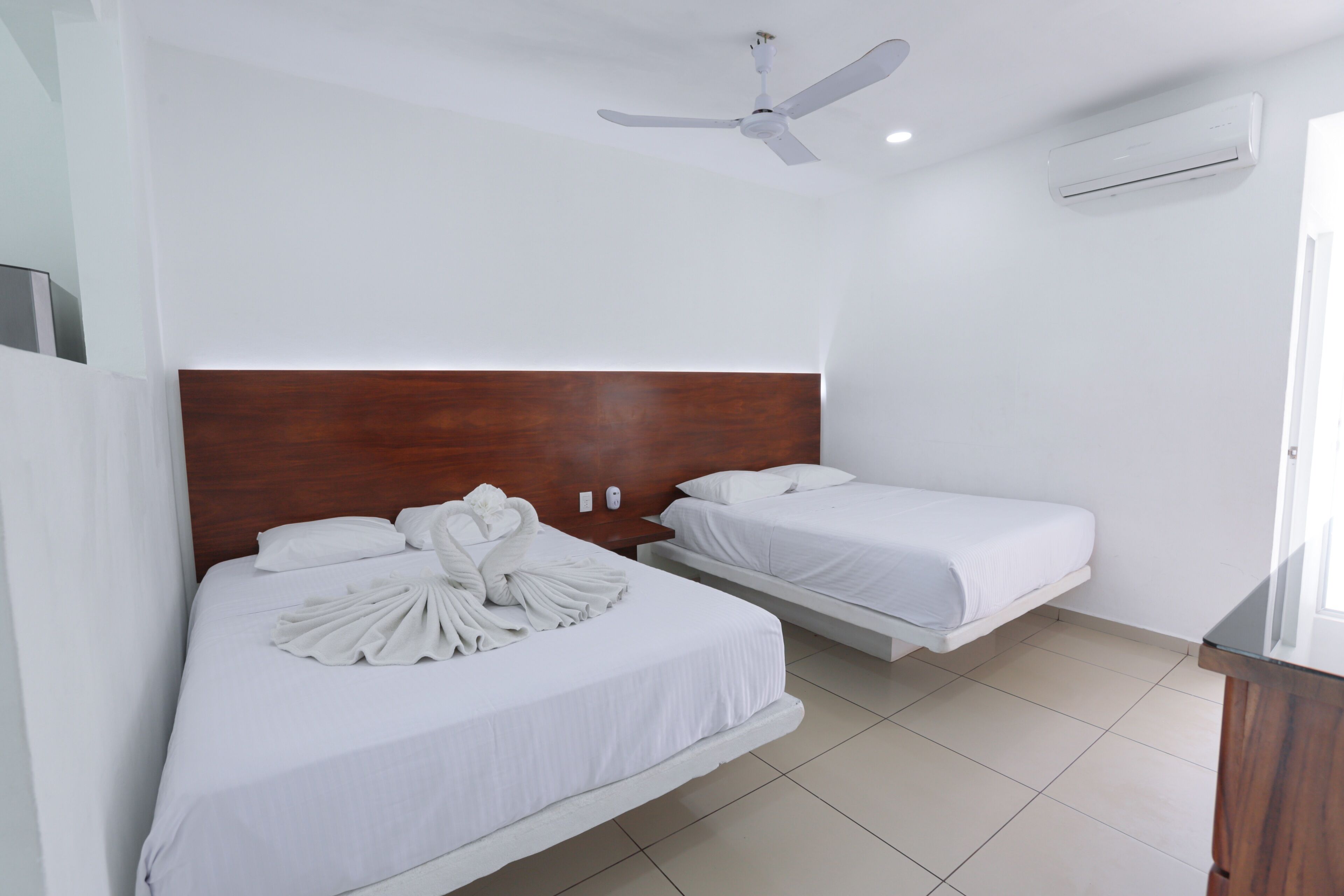 A hotel room with two beds, a ceiling fan, an air conditioning unit, and a wooden headboard.