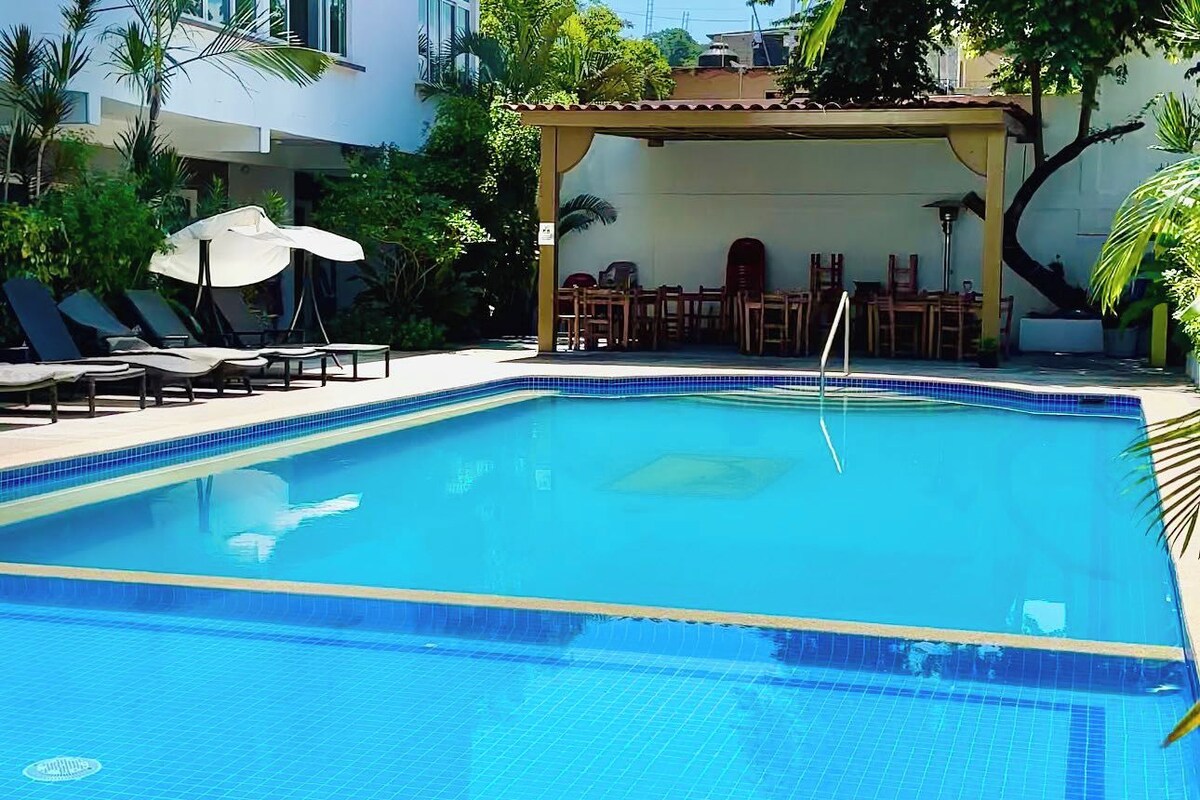 Una piscina rectangular con azulejos azules, rodeada de sillones de playa y una zona de estar cubierta con mesas y sillas.