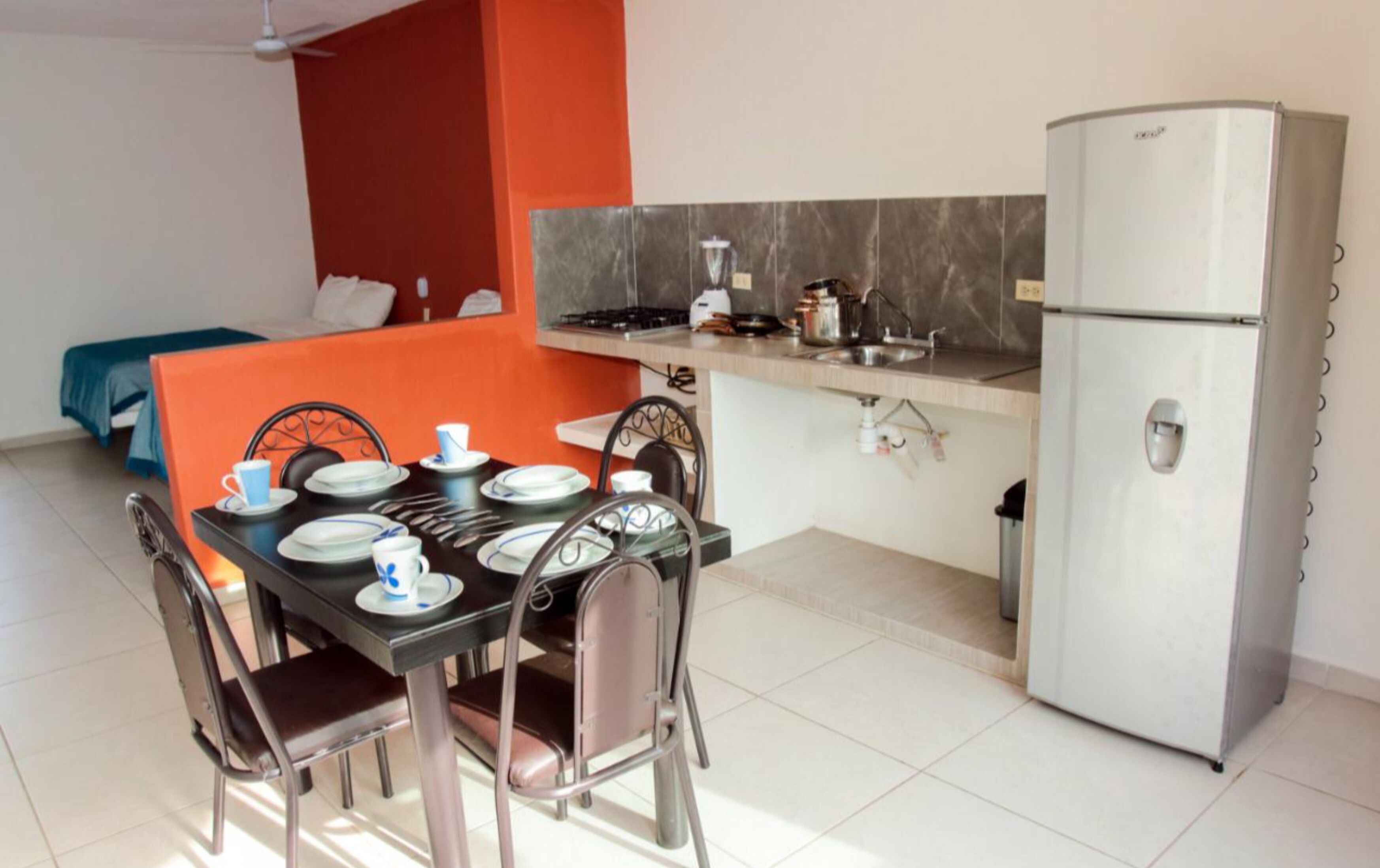A compact kitchen with a refrigerator, a dining table set with plates, cups, and cutlery, and a bed in the background.