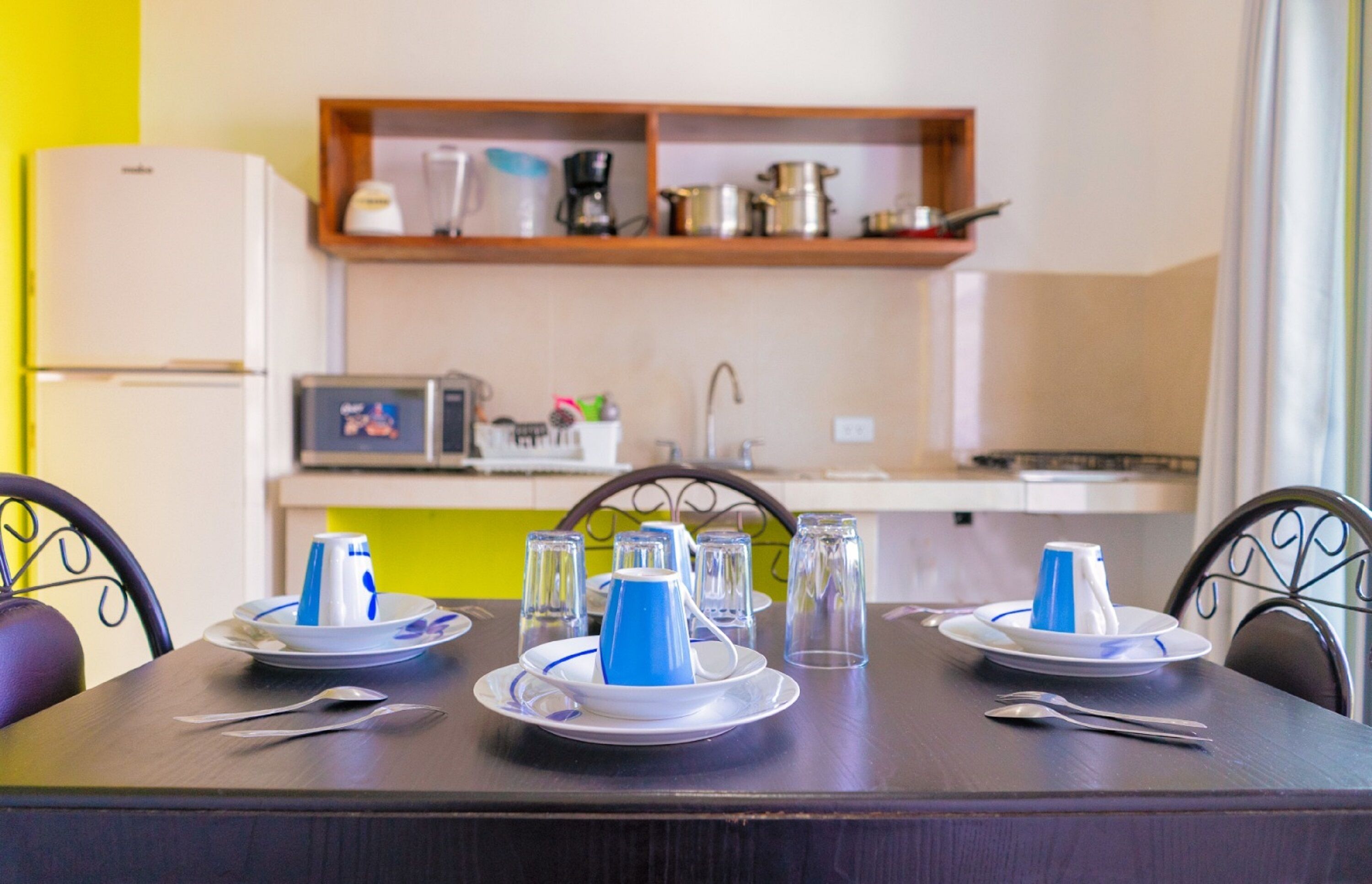 A dining table set with plates, cups, and cutlery in a room with a kitchen in the background.