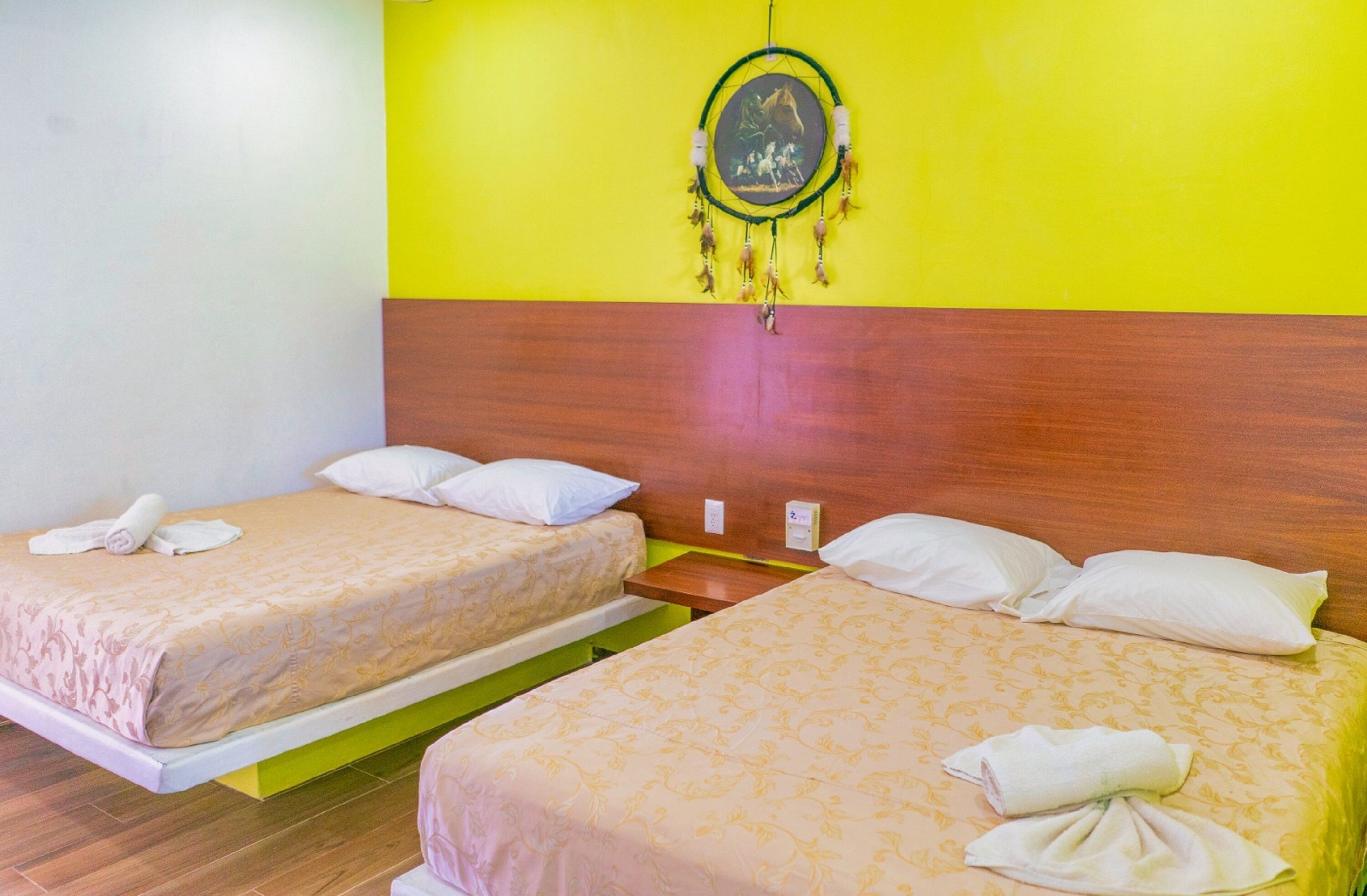 A hotel room with two single beds, a wooden headboard, a wall-mounted dreamcatcher, and a small bedside table with a lamp.