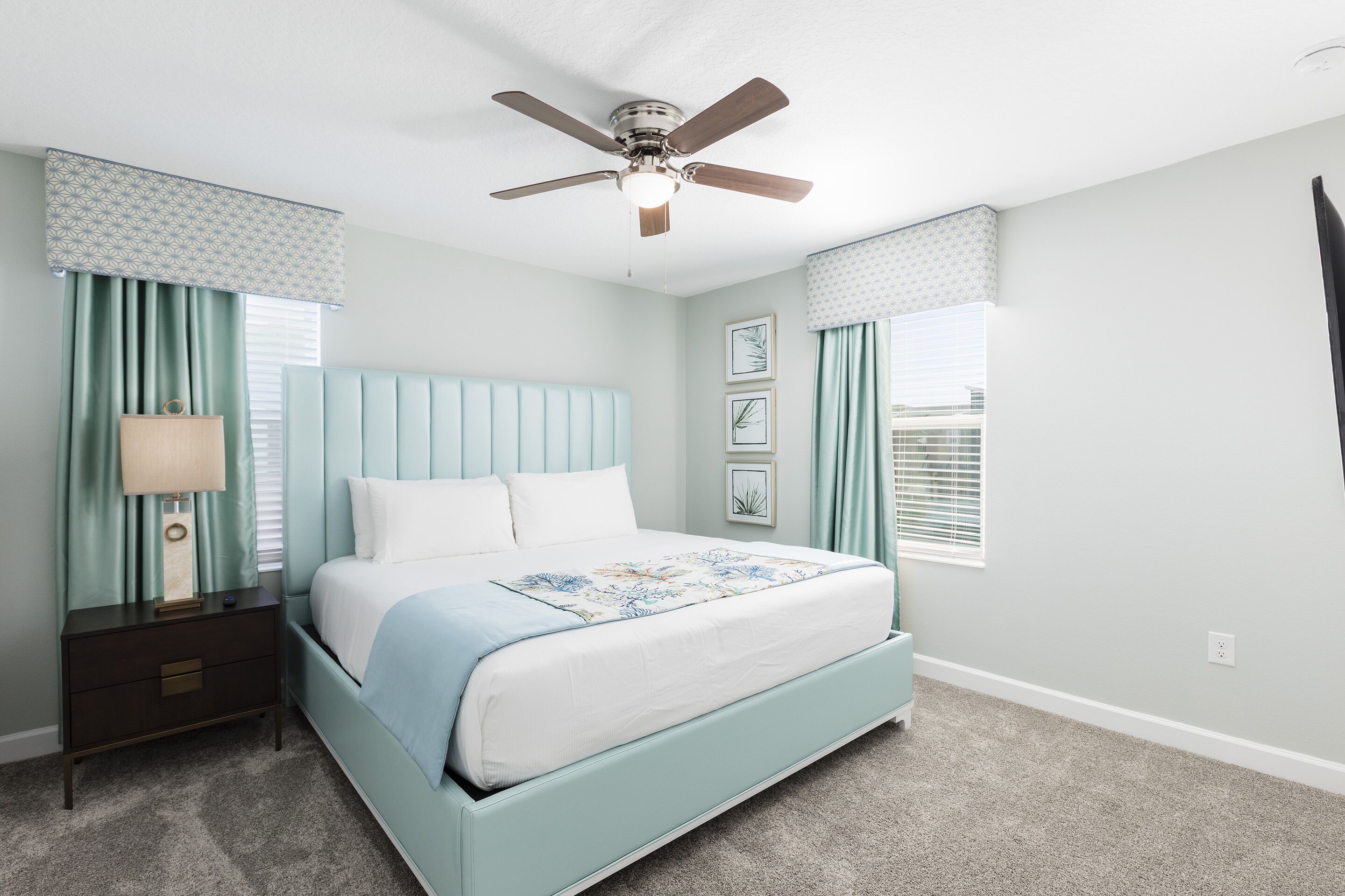 A bedroom with a bed, nightstand, lamp, ceiling fan, and two windows with curtains.
