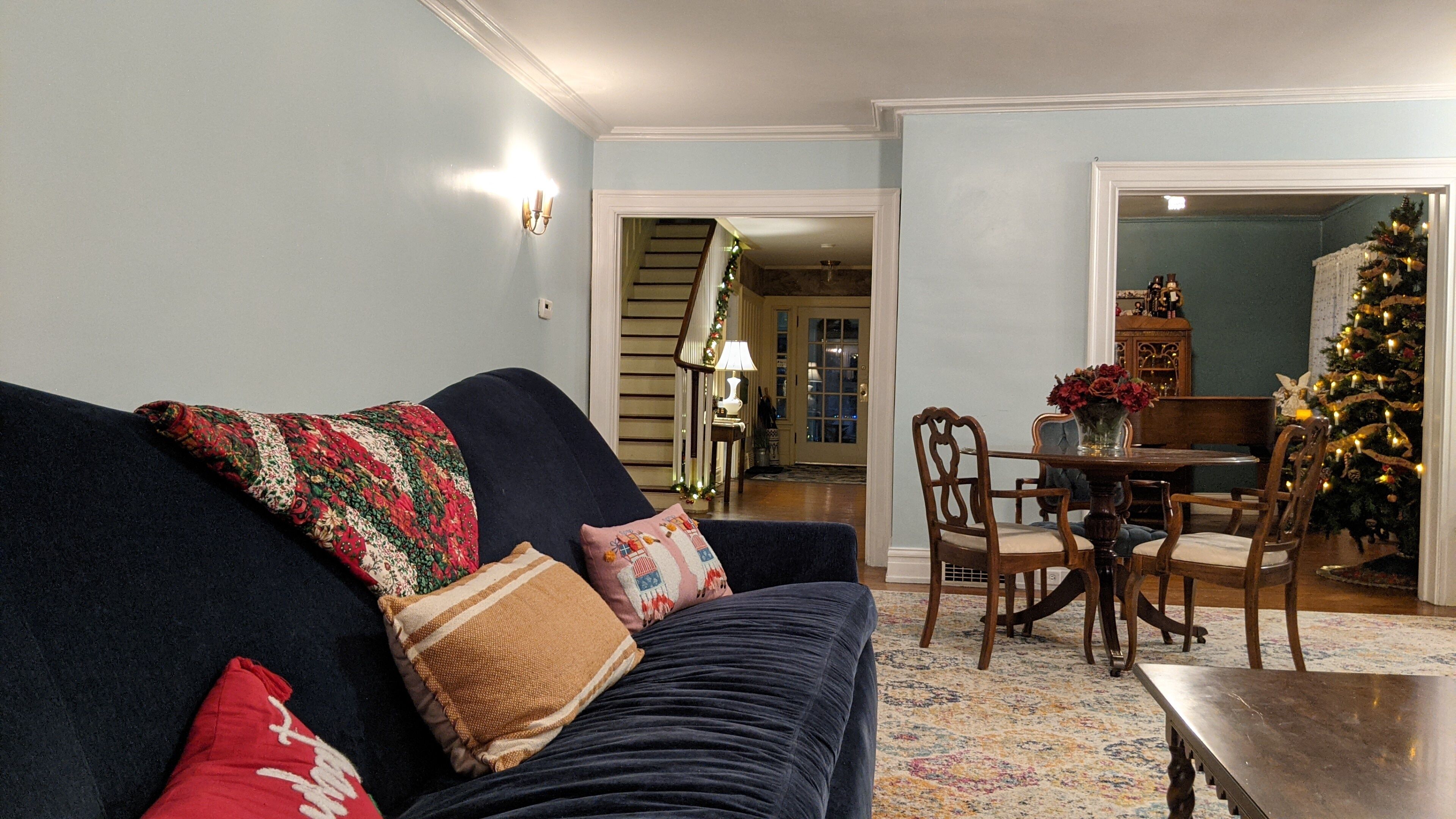A living room with a dark blue sofa, a wooden dining table with chairs, a Christmas tree, and a staircase leading to another room.