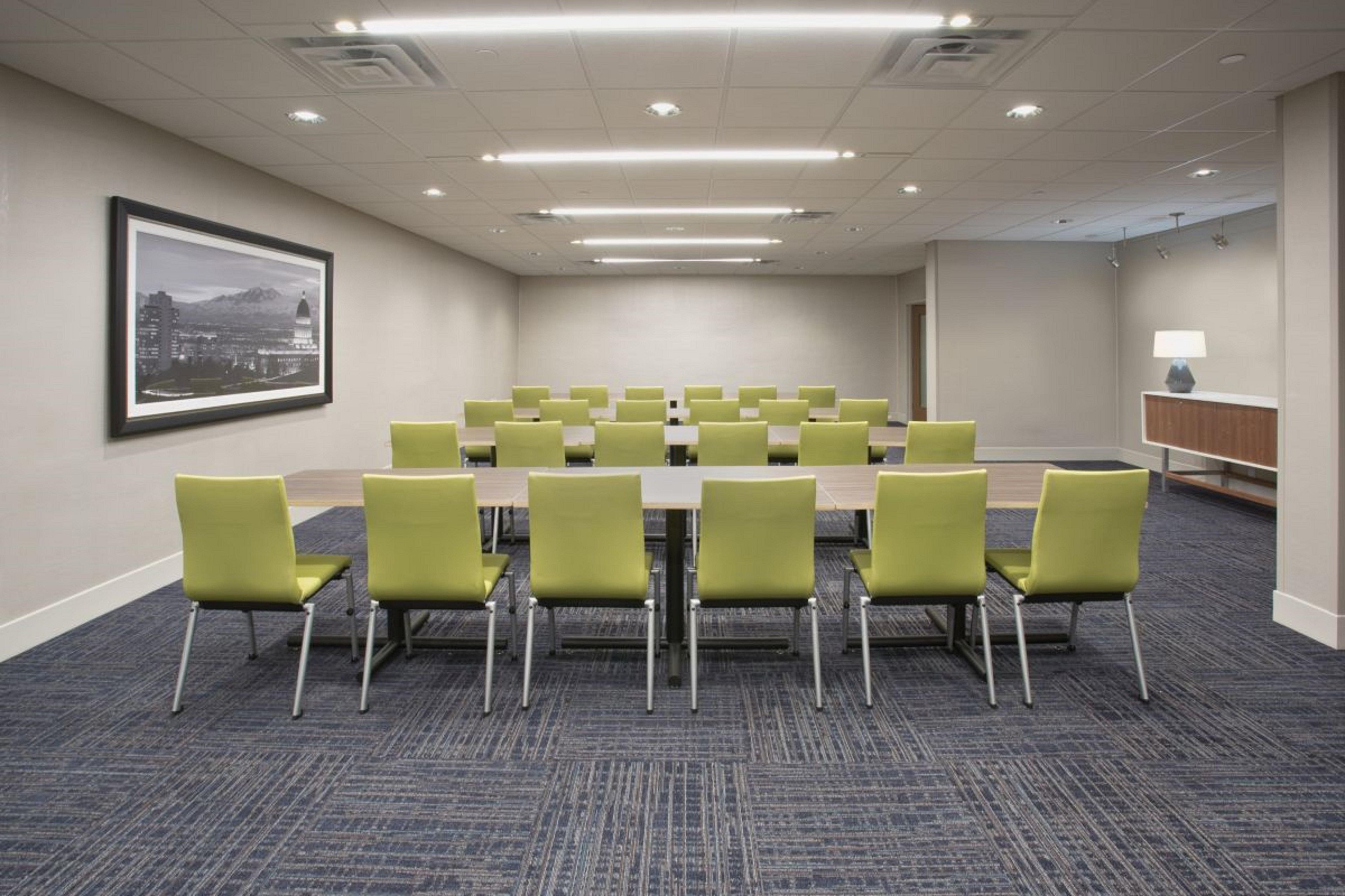 A conference room with a long table, green chairs, carpeted floor, and a framed cityscape picture on the wall.
