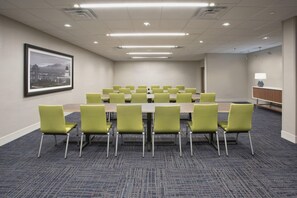 Una sala de conferències amb una taula llarga, caires verds, sòl amb moqueta i una imatge emmarcada d'un paisatge urbà a la paret.