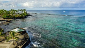 A coastal view with clear turquoise waters, a rocky shoreline, and a small beach area with palm trees and a few buildings. - Kahalu’u Surf and Snorkel Beachfront Paradise, with Hot Tub (Kailua-Kona)