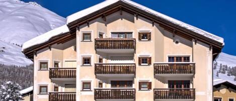 A multi-story building with balconies, set against a snowy mountain backdrop.