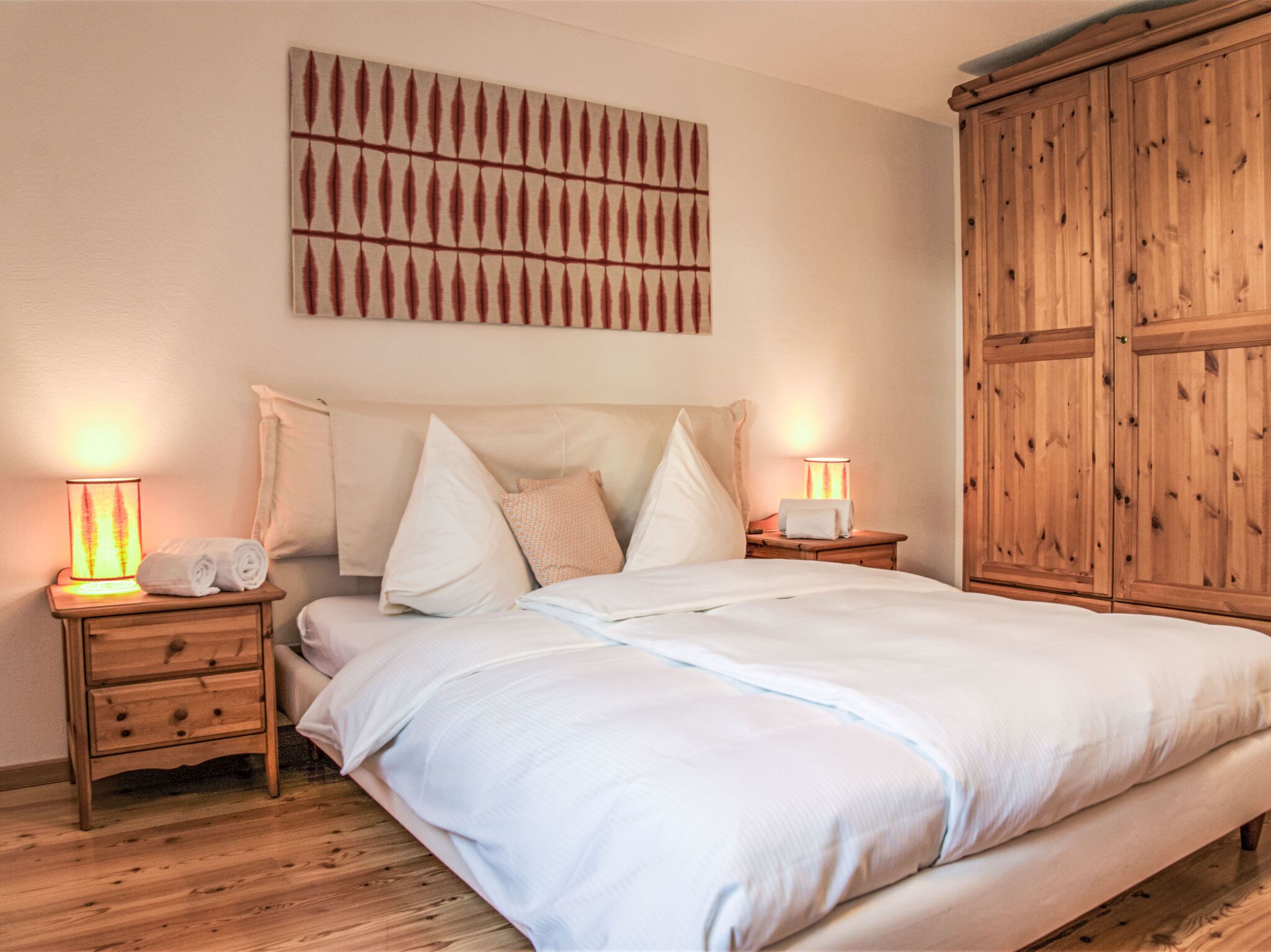 Ein Schlafzimmer mit einem großen Bett, zwei Nachttischen inklusive Lampen, einem Holzschrank und einem an der Wand befestigten Kunstwerk.