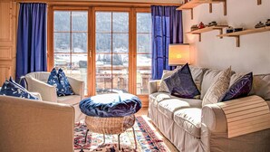 A cozy living room with a sofa, armchair, and ottoman, large windows with curtains, and wooden shelves with books and decorative items.