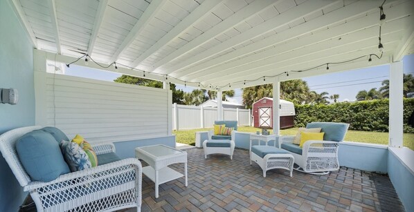 A covered patio area with wicker furniture, a small table, and a red shed in the background. - Beautiful, Private Beach House - 1 Block To The Beach - AC 3 Bedroom - Sleeps 8 (Cocoa Beach)