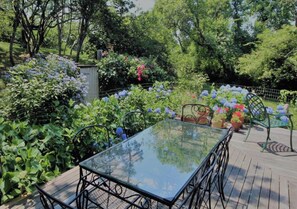 An outdoor patio with a glass-top table and metal chairs, surrounded by lush greenery and flowering plants. - Westside Story...an enchanting island getaway. (New Shoreham)