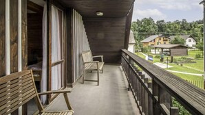 Un balcon en bois avec deux chaises en bois, un sol en béton et une vue sur des maisons et de la verdure en arrière-plan.