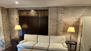 A cozy living room with a sofa, two side tables with lamps, a woven ottoman, and a brick wall with decorative panels. - Cozy Furnished Apartment (New Orleans)