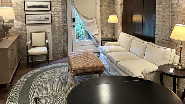 A modern living room with a sofa, a dining table, and a chair. There is a door with a view to the outside, a lamp, and a wall with a decorative panel. - Cozy Furnished Apartment (New Orleans)