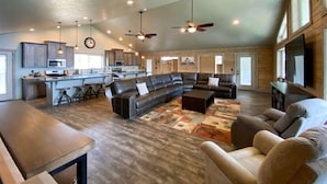 A spacious living area with a large L-shaped sectional sofa, a coffee table, and a dining table. There is a kitchen area with a bar and stools, and a television mounted on the wall.