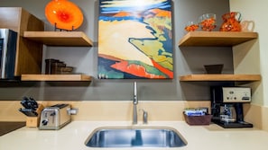 Una cocina con diseño moderno, que incluye un lavamanos de acero inoxidable, una cafetera y un tostador. Hay un cuadro sobre el lavamanos y una estantería de madera con objetos decorativos.