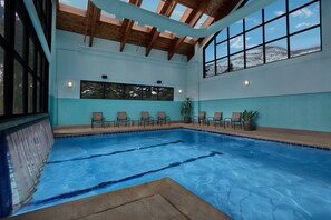 An indoor swimming pool with clear blue water, wooden beams on the ceiling, and a row of chairs along the poolside.