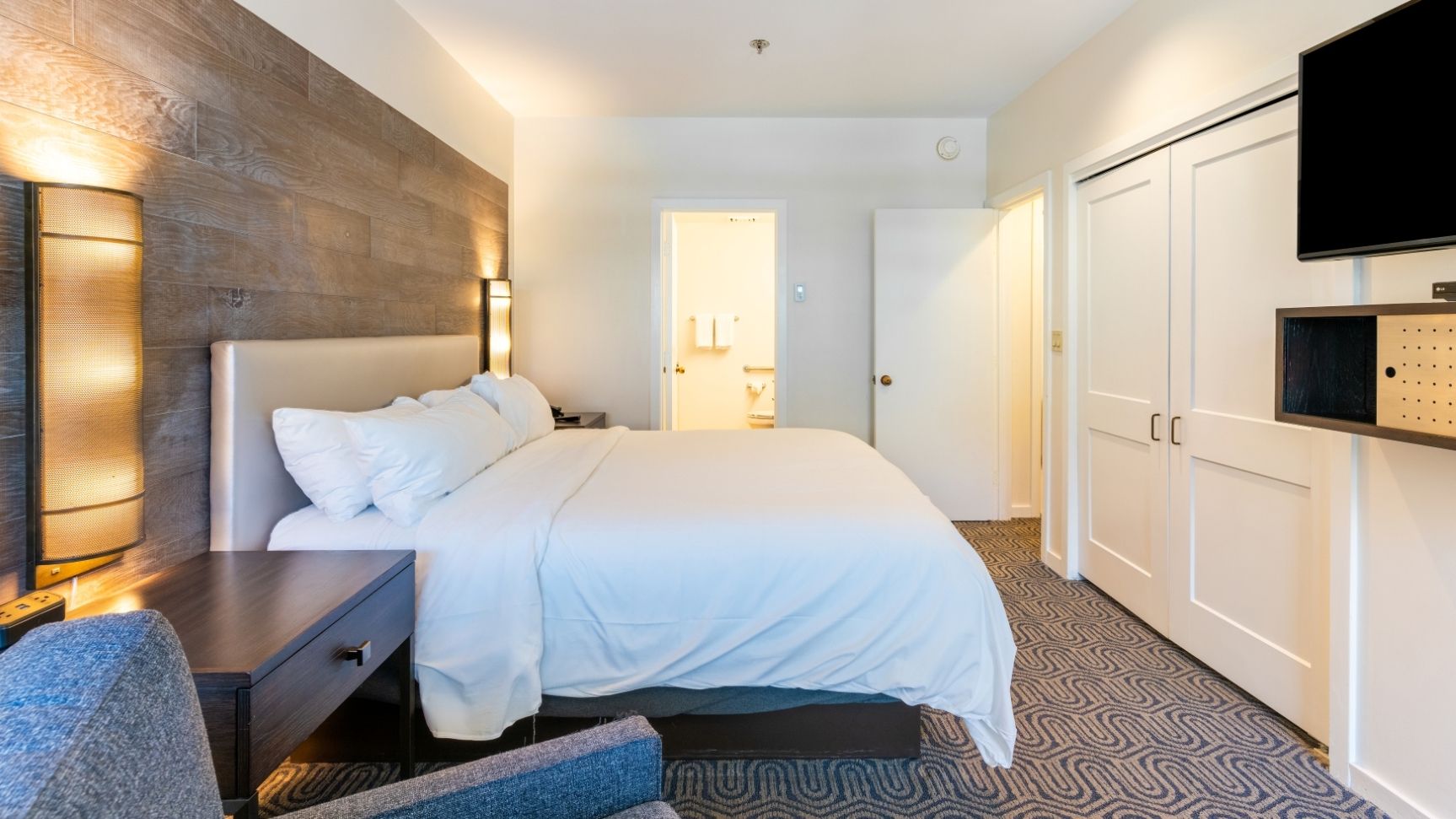 Quarto de hotel com cama, mesa de cabeceira, luminária, guarda-roupa e televisão.