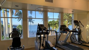Un gimnasio con equipo de cardio, amplias ventanas y vista a palmeras y al agua.