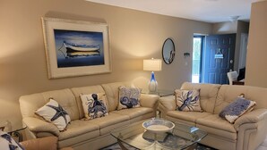 Een woonkamer met een beige hoekbank, een glazen salontafel en decoratieve kussens met een patroon. Op de muur hangt een lijst met een afbeelding van een boot, en er staat een lamp en een deur met een nummerbord.