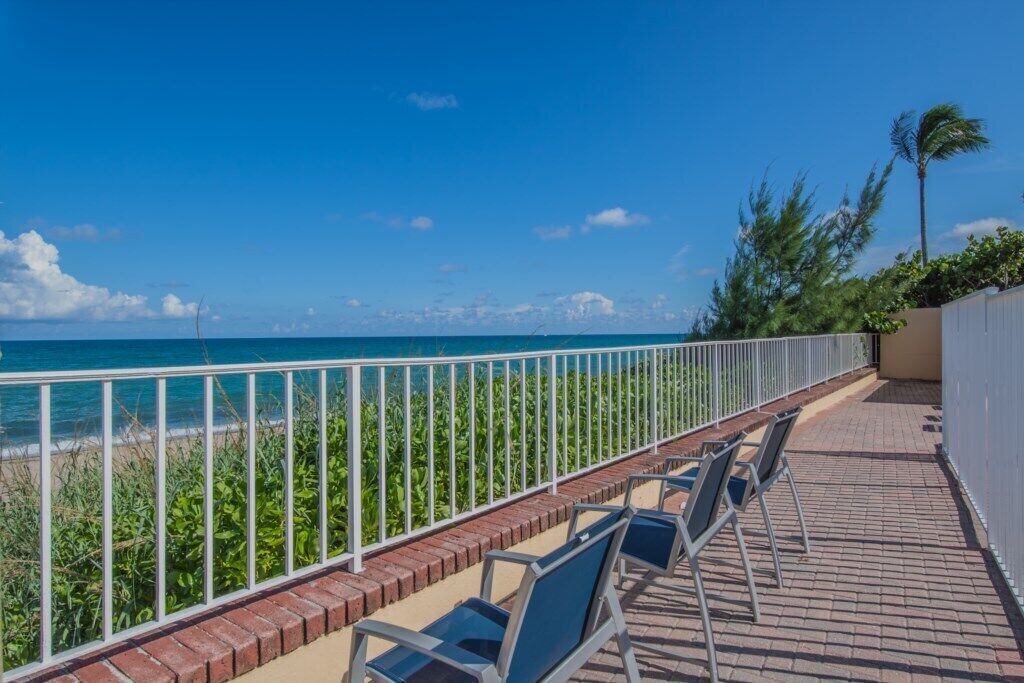 A balcony with chairs overlooking a beach, a white fence, and a palm tree.