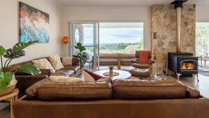 A modern living room with a stone fireplace, leather sofas, and a large window offering a view of the outdoors. - Summerville Treetops: A Stunning Valley Retreat (Yallingup Siding)