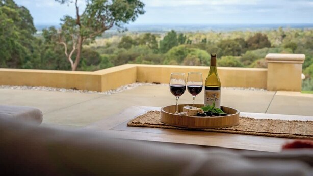 A terrace with a table set for two, featuring a bottle of wine and glasses, overlooking a landscape with trees and a distant view. - Summerville Treetops: A Stunning Valley Retreat (Yallingup Siding)