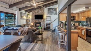 A spacious living area with a stone fireplace, a large TV mounted above it, and a cozy seating area with a sofa and a dining table. - Ski-In/Out Remodeled Mountain View Penthouse 1025 (Steamboat Springs)