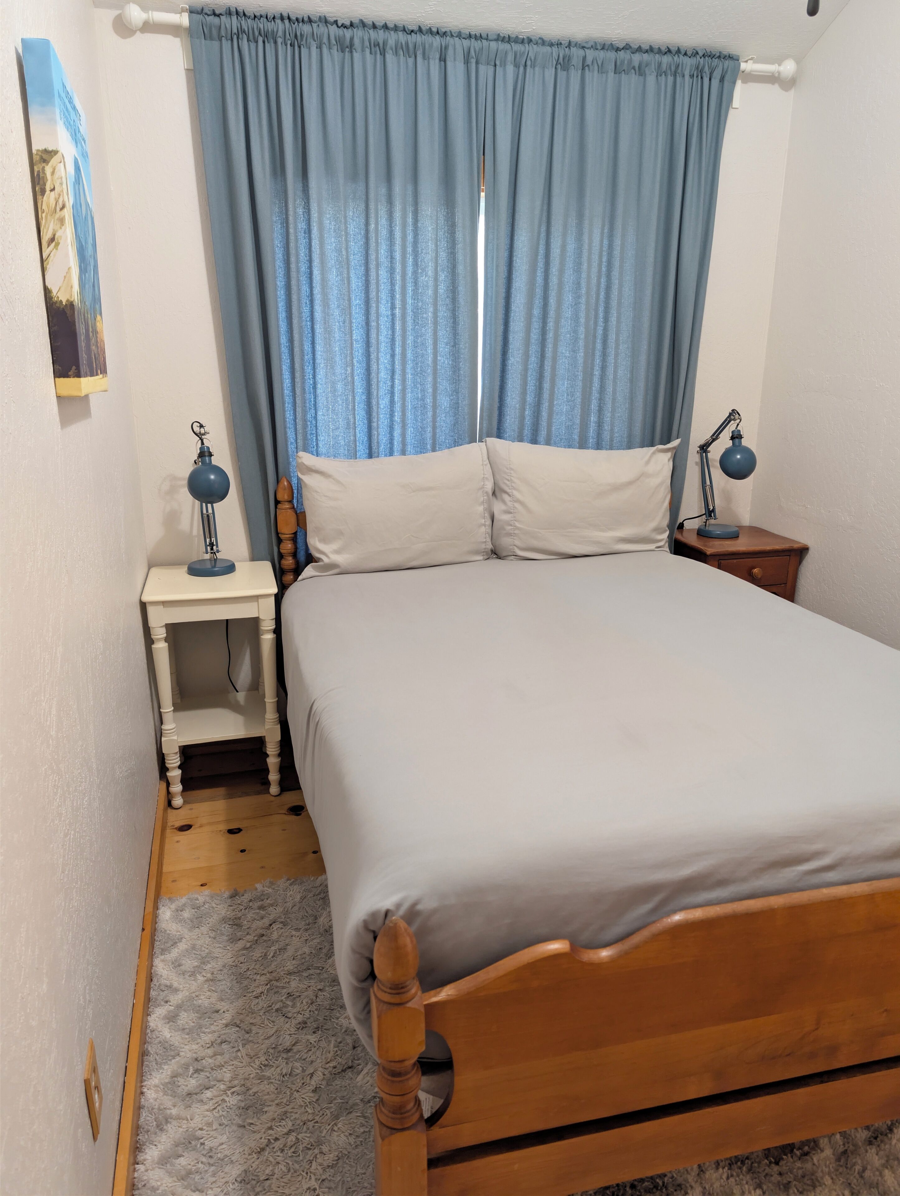 A bedroom with a wooden bed, two bedside tables with lamps, a blue curtain, and a painting on the wall.
