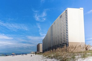 Um hotel moderno de vários andares, com design arquitetônico contemporâneo, situado ao lado de uma praia com areia visível e vegetação rala.