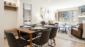 A modern dining area with a wooden table set for four, surrounded by black upholstered chairs. A grey sofa and a small coffee table are visible in the background.