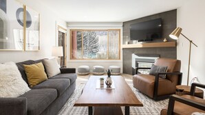 A modern living room with a grey sofa, a brown leather armchair, a wooden coffee table, and a large TV mounted on the wall.