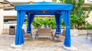 A covered outdoor seating area with a stone floor, surrounded by greenery and a waste bin. - Waikiki | Waikiki Beach Stay | Pool & Hot Tub | 2BR (Honolulu, Ohau)