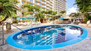 Una piscina con sillas de descanso y sombrillas, rodeada de palmeras y un edificio de varios pisos.