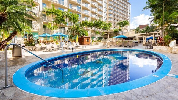 Ein Swimmingpool mit Liegestühlen und Sonnenschirmen, umgeben von Palmen und einem mehrstöckigen Gebäude.