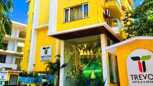Un edificio de hotel amarillo con entrada de vidrio, palmeras y un letrero que dice Trevon Hotels & Resorts.
