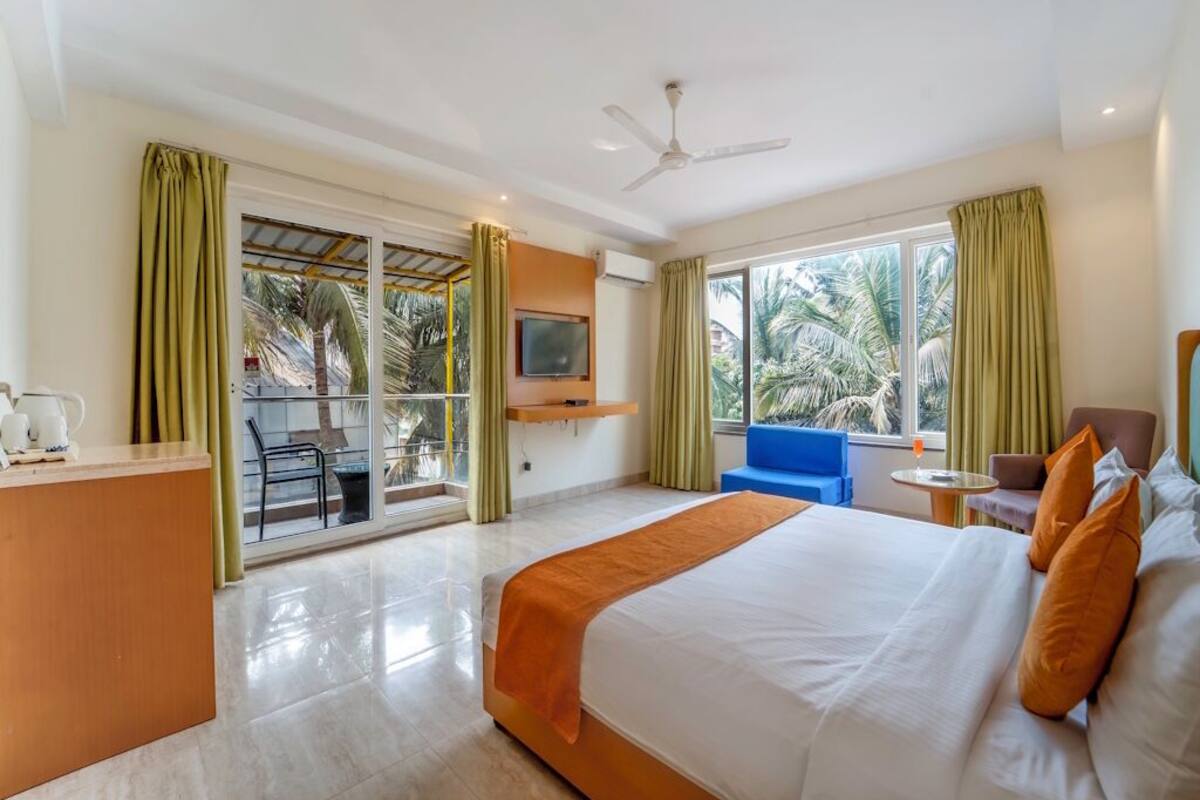 Quarto de hotel com cama, escrivaninha, TV e varanda com vista para palmeiras.
