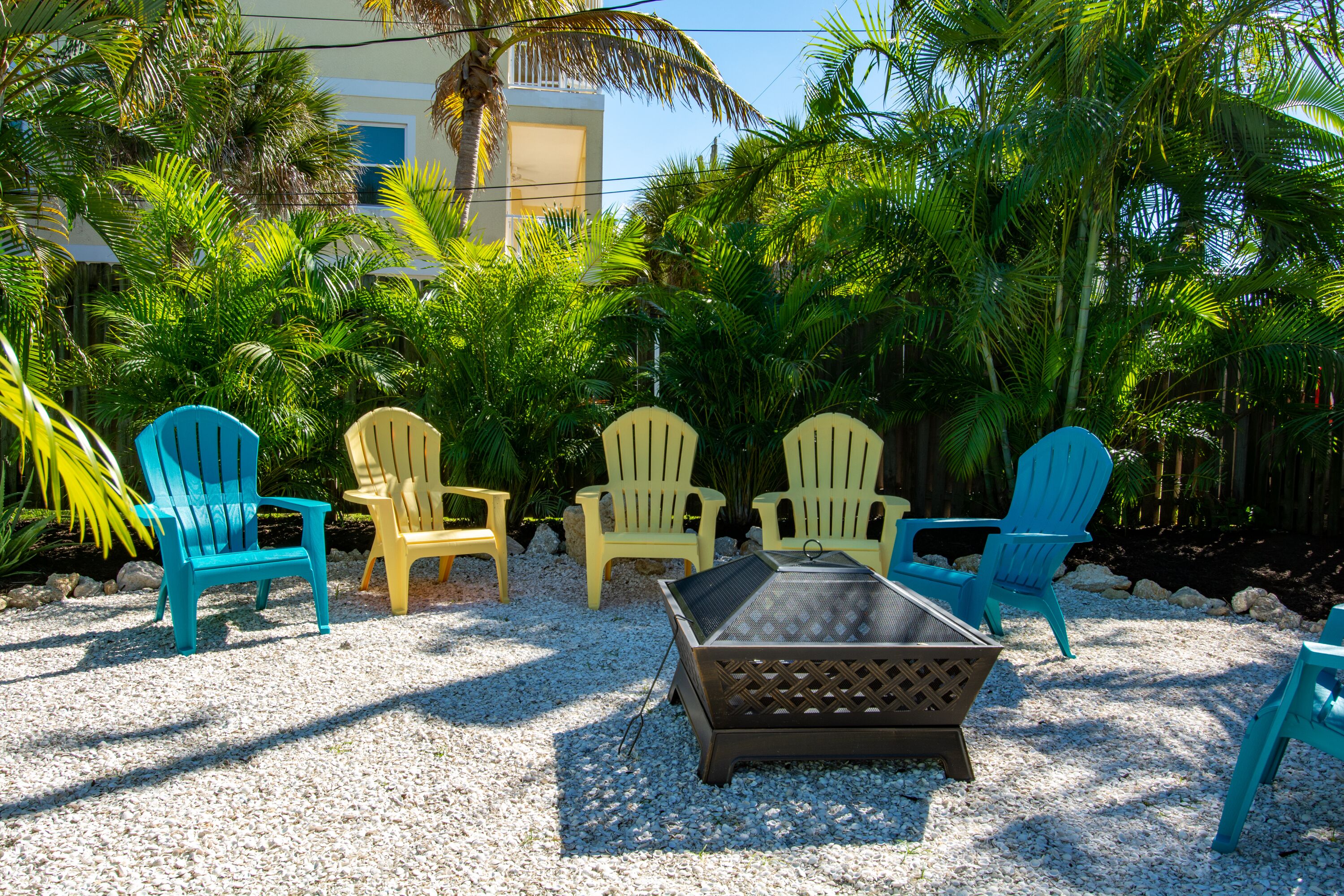 A patio area with outdoor seating including chairs and a table, surrounded by lush greenery and a building in the background.