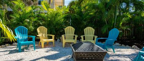 A patio area with outdoor seating including chairs and a table, surrounded by lush greenery and a building in the background.