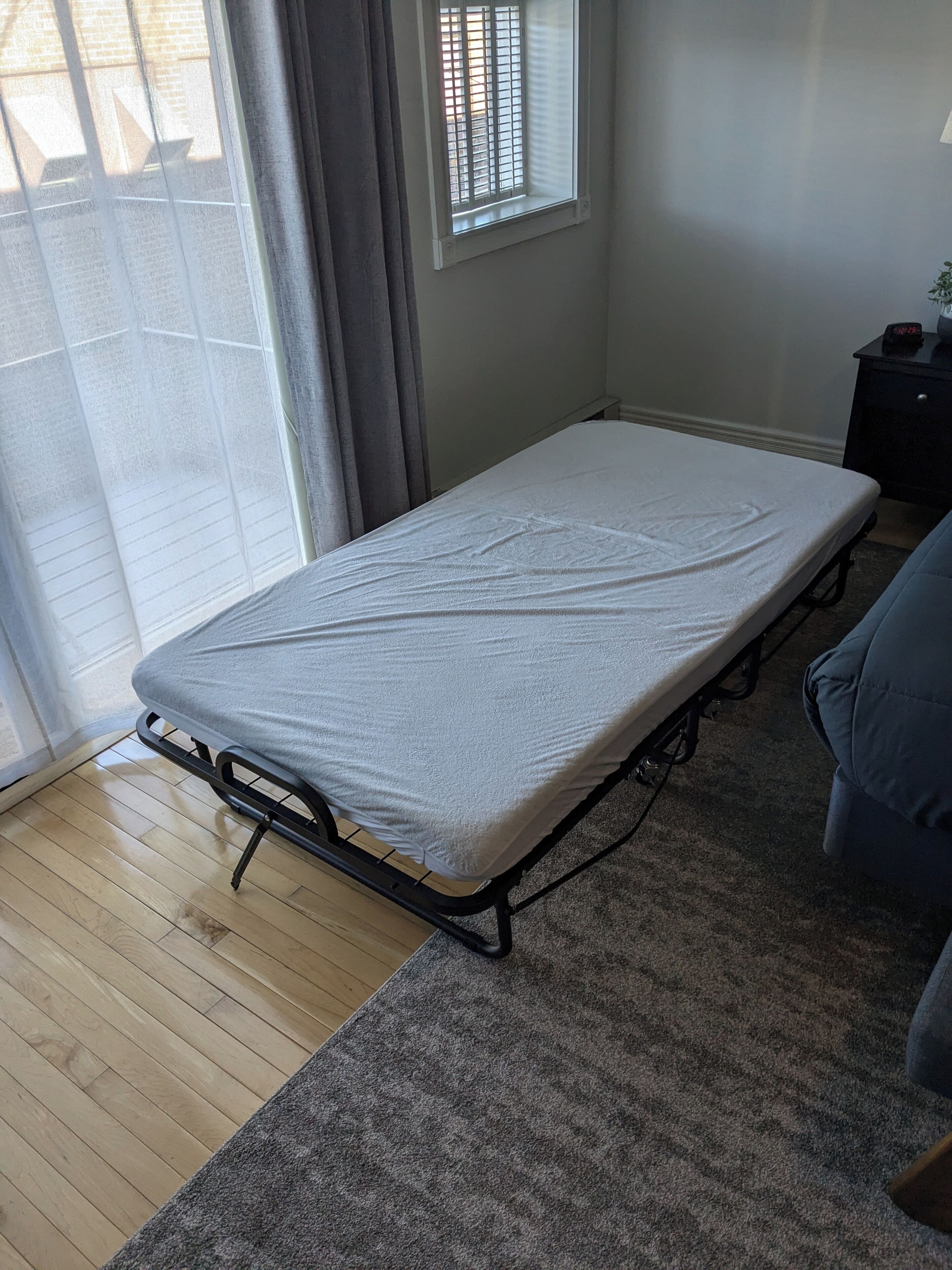 A room with a single bed, a small table, a chair, a window with blinds, and a grey carpet.