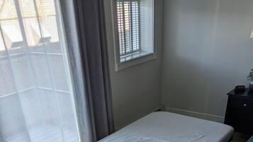 A room with a single bed, a small table, a chair, a window with blinds, and a grey carpet.