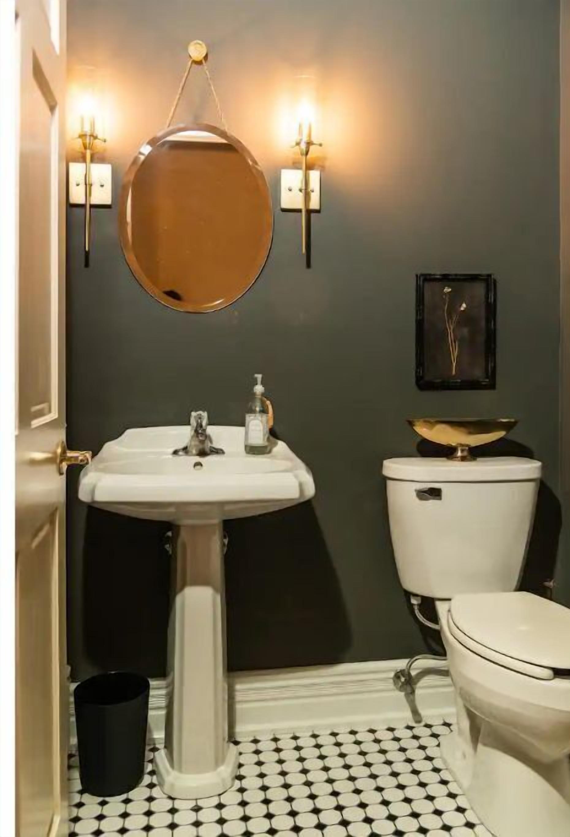 Un baño con un lavamanos blanco, un inodoro y un espejo redondo. Hay apliques con luces, un cesto de basura negro y un cuadro en la pared.