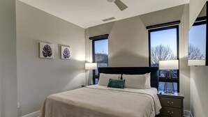 A bedroom with a bed, two windows, a ceiling fan, and wall art. - Biltmore Avenue Balcony Condo (Asheville)