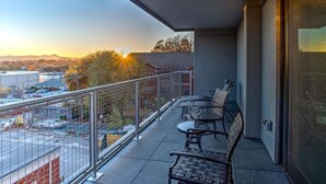 A balcony with a view of a parking lot and buildings, furnished with metal chairs and a table.